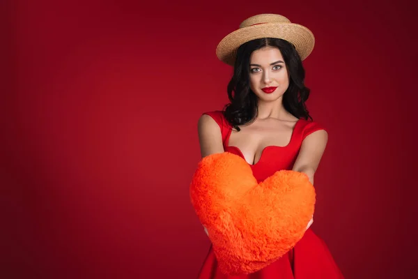 Menina atraente em vestido vermelho mostrando travesseiro em forma de coração isolado no vermelho — Fotografia de Stock