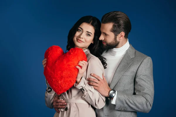 Girlfriend holding heart shaped pillow and boyfriend hugging her isolated on blue — Stock Photo