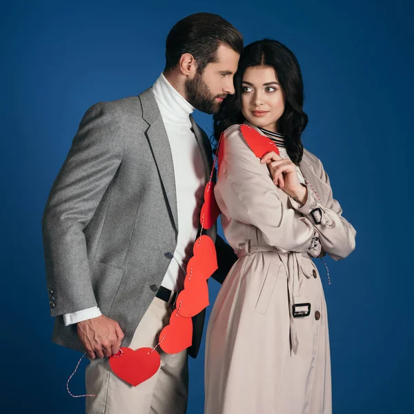 Atractiva pareja sosteniendo guirnalda de corazones rojos aislados en azul - foto de stock