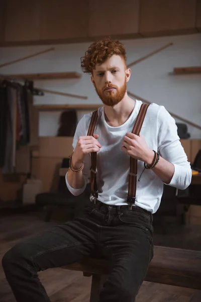 Guapo elegante joven diseñador de moda en tirantes sentado y mirando a la cámara - foto de stock