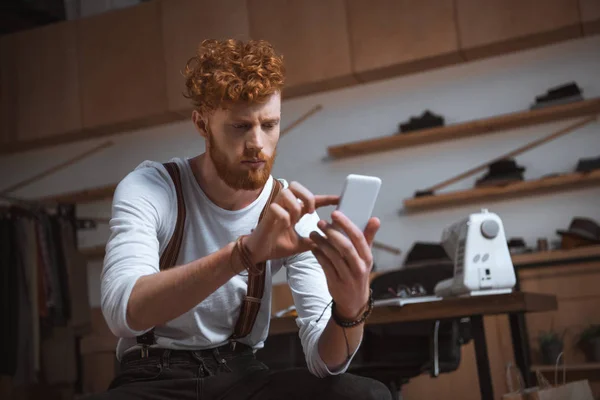 Enfocado joven diseñador de moda masculino usando teléfono inteligente mientras está sentado en el taller - foto de stock