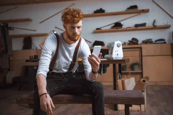Diseñador de moda joven serio sosteniendo el teléfono inteligente y mirando a la cámara en el taller - foto de stock