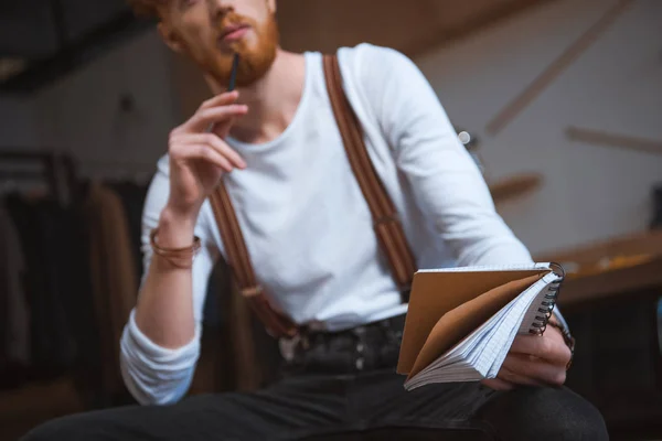 Corte tiro de jovem designer de moda masculina com notebook e lápis sentado no local de trabalho — Fotografia de Stock