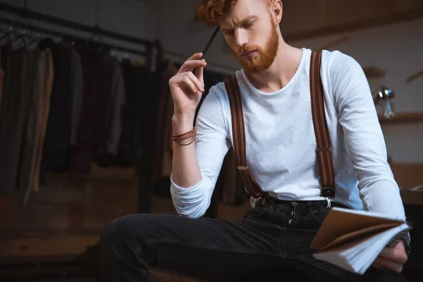 Pensativo joven diseñador de moda masculino con cuaderno y lápiz sentado en el lugar de trabajo - foto de stock