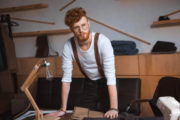 Красивый молодой дизайнер в очках, склоняющийся за столом с швейными инструментами в мастерской — стоковое фото