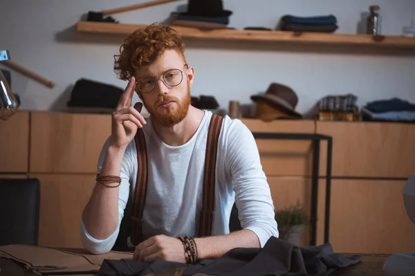 Bonito jovem designer de moda em óculos olhando para a câmera enquanto sentado no local de trabalho — Fotografia de Stock