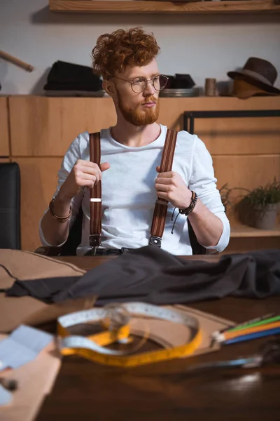 Nachdenkliche junge bärtige Modedesignerin mit Brille, die am Arbeitsplatz wegschaut — Stockfoto