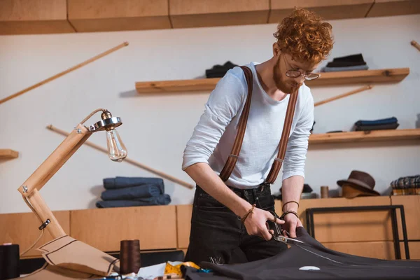 Молодой дизайнер в очках, работающий с ножницами и тканями в мастерской — стоковое фото