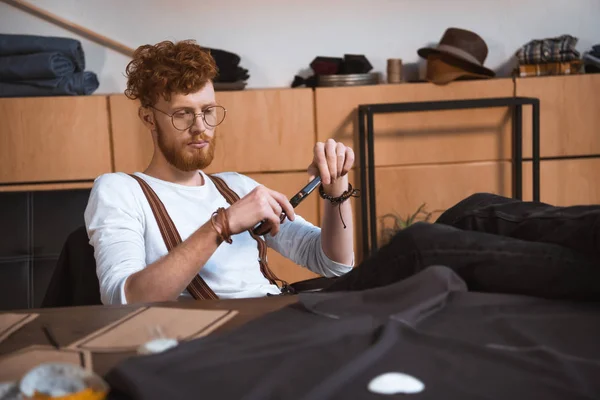 Joven diseñador de moda masculino sosteniendo tijeras mientras está sentado en la mesa con patrones de costura y tela - foto de stock