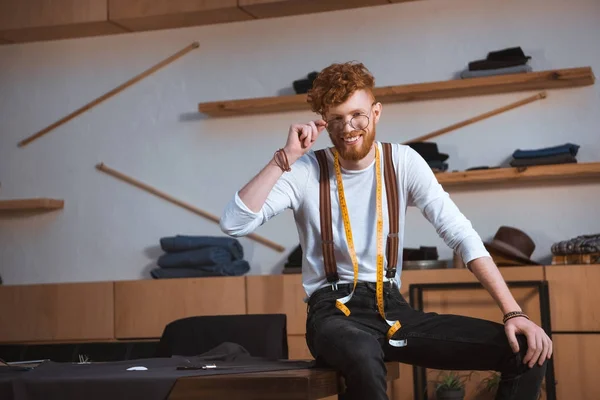 Junger männlicher Modedesigner mit Brille und Maßband um den Hals lächelt am Arbeitsplatz in die Kamera — Stockfoto
