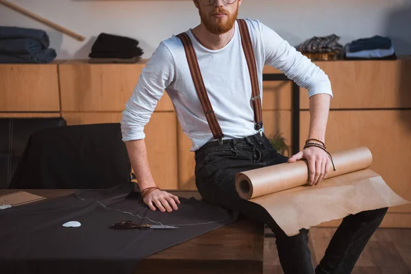 Tiro recortado de joven diseñador de moda masculino sentado en la mesa con la tela y herramientas de costura en el taller - foto de stock