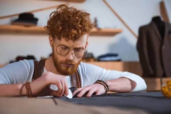 Konzentrierte junge bärtige Modedesignerin arbeitet in Werkstatt mit Stoff — Stockfoto