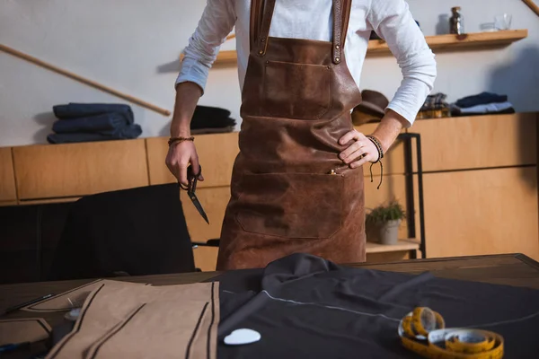 Cropped shot of male fashion designer in apron working with sewing tools and fabric at workshop — Stock Photo