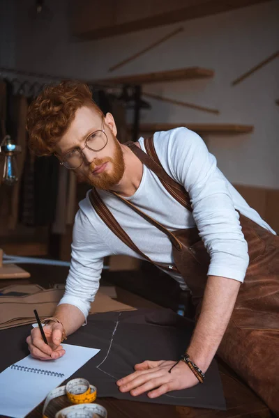 Handsome young fashion designer in apron and eyeglasses taking notes in notebook — Stock Photo
