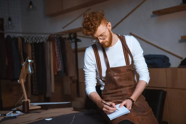 Giovane stilista di moda maschile in grembiule e occhiali prendere appunti nel quaderno — Foto stock