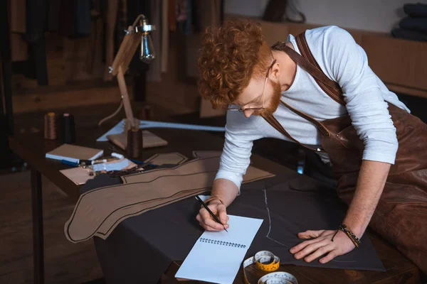 Jeune créateur de mode masculin dans le tablier et les lunettes prendre des notes dans le carnet — Photo de stock