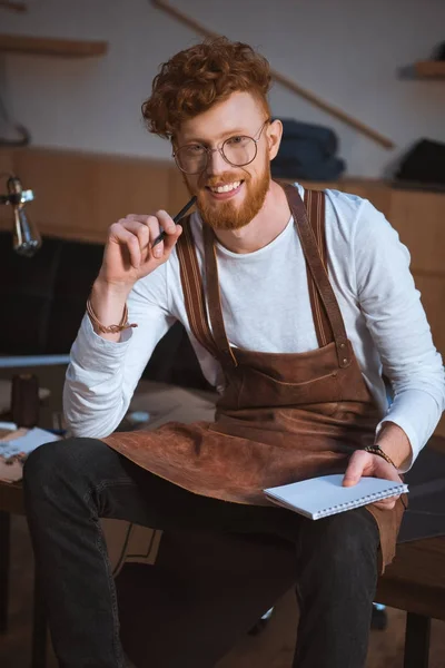 Handsome young fashion designer in eyeglasses holding pencil with notebook and smiling at camera — Stock Photo