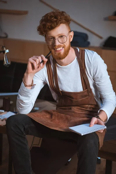 Bonito jovem designer de moda em óculos segurando lápis com notebook e sorrindo para a câmera — Fotografia de Stock