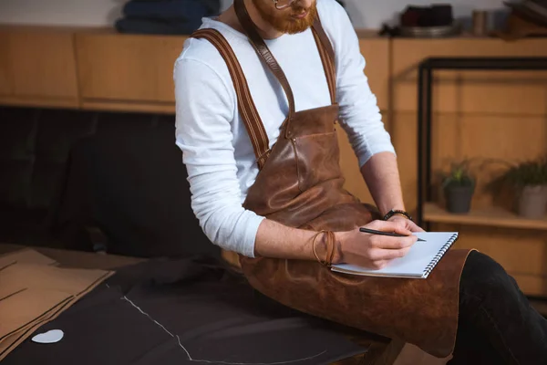 Cropped shot of bearded male fashion designer in apron taking notes in notebook — Stock Photo