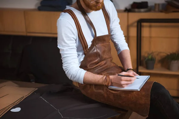 Plan recadré du créateur de mode masculin barbu dans tablier prenant des notes dans le carnet — Photo de stock