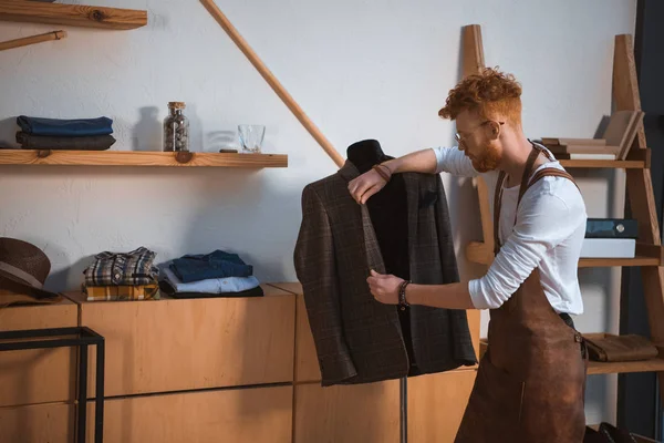 Giovane stilista di moda maschile in grembiule e occhiali guardando giacca vestito sul manichino in officina — Foto stock