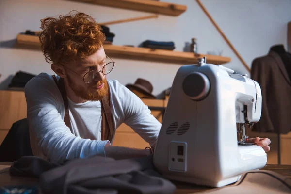 Enfocado joven diseñador de moda en gafas que trabajan con la máquina de coser - foto de stock