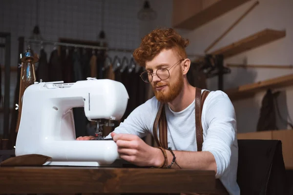 Sorrindo jovem designer de moda em óculos trabalhando com máquina de costura — Fotografia de Stock