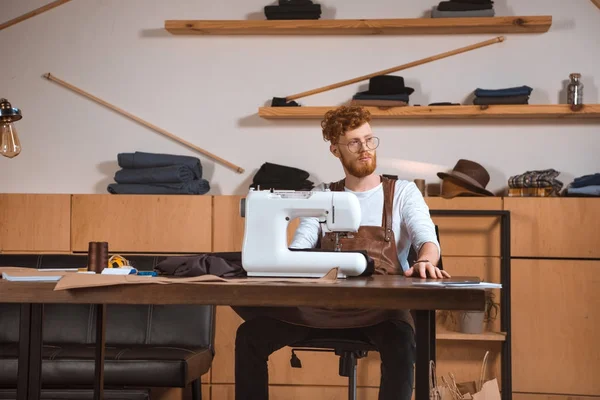 Guapo joven diseñador de moda en gafas mirando hacia otro lado mientras trabaja con la máquina de coser en el taller - foto de stock