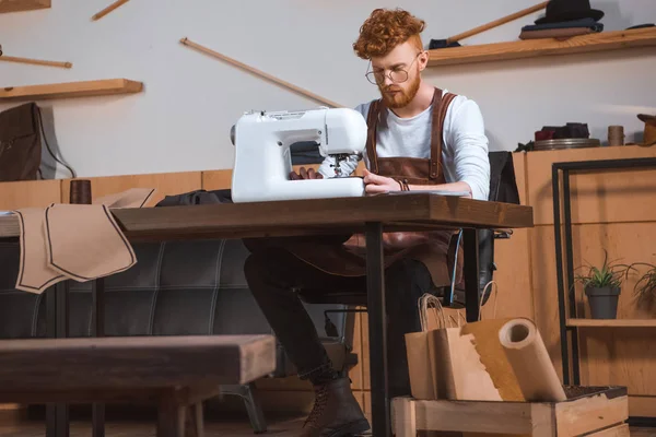 Jovem designer de moda em avental e óculos trabalhando com máquina de costura na oficina — Fotografia de Stock