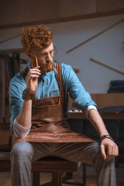 Jeune créateur de mode masculin coûteux dans tablier et lunettes assis et pensant avec crayon à la main — Photo de stock