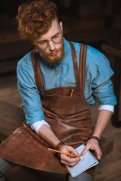 Junger männlicher Modedesigner in Schürze und Brille notiert im Notizbuch — Stockfoto