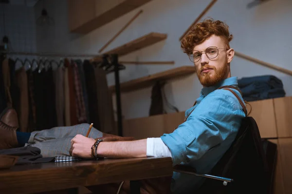 Junger männlicher Modedesigner mit Brille blickt in die Kamera, während er am Arbeitsplatz Notizen macht — Stockfoto