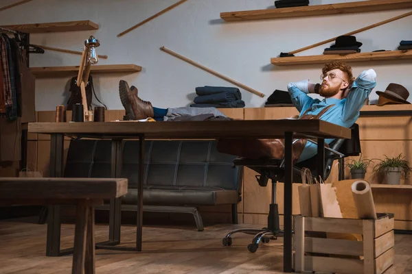 Jeune créateur de mode masculin dans tablier et lunettes assis avec les mains derrière la tête sur le lieu de travail — Photo de stock