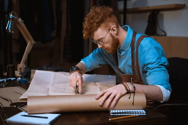 Joven diseñador de moda masculino en delantal y anteojos haciendo patrones de costura en el lugar de trabajo - foto de stock