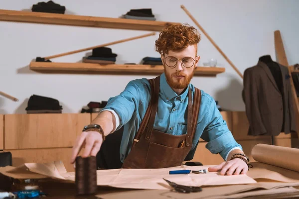 Jovem designer de moda masculina em avental e óculos sentado no local de trabalho — Fotografia de Stock