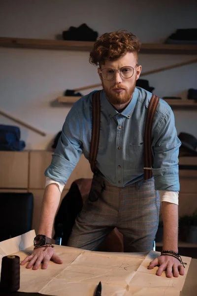 Guapo joven diseñador de moda en gafas mirando a la cámara mientras se inclina en la mesa con papel artesanal - foto de stock