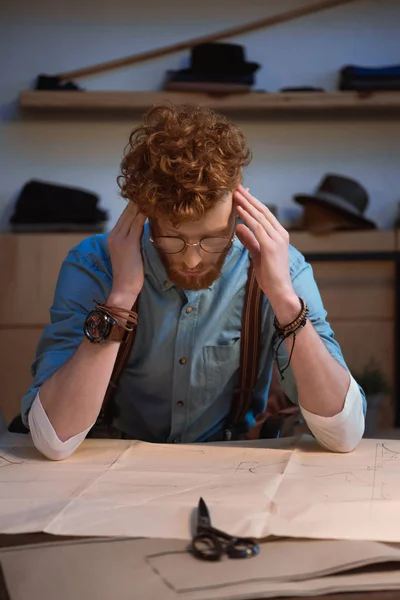 Tired young fashion designer in eyeglasses sitting at table with craft paper and scissors — Stock Photo