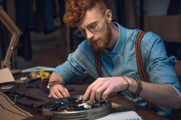 Jovem designer de moda masculina em óculos trabalhando com botões coloridos e tecido na oficina — Fotografia de Stock