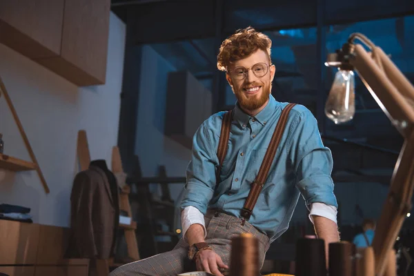 Jeune créateur de mode souriant dans des lunettes assis sur la table sur le lieu de travail — Photo de stock