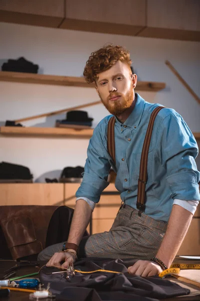 Handsome young fashion designer looking at camera while sitting on table at workplace — Stock Photo
