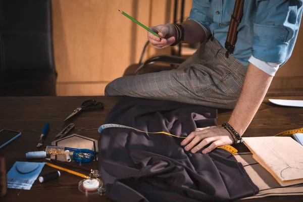 Cropped shot of young fashion designer holding pencil and sitting on table at workplace — Stock Photo