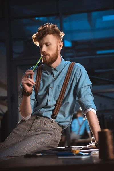 Stylish young fashion designer in suspenders sitting on table at workplace — Stock Photo