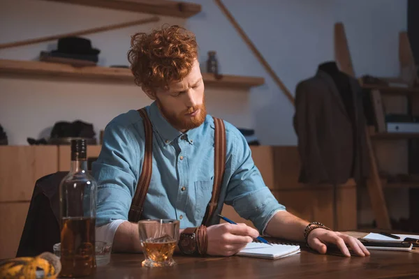 Jeune styliste prenant des notes et buvant du whisky sur le lieu de travail — Photo de stock