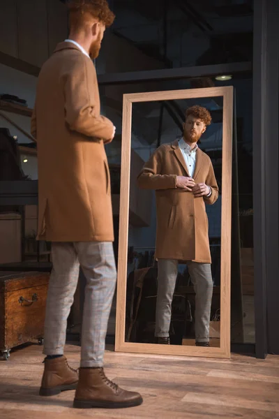 Stylish young man wearing overcoat and looking at mirror in boutique — Stock Photo