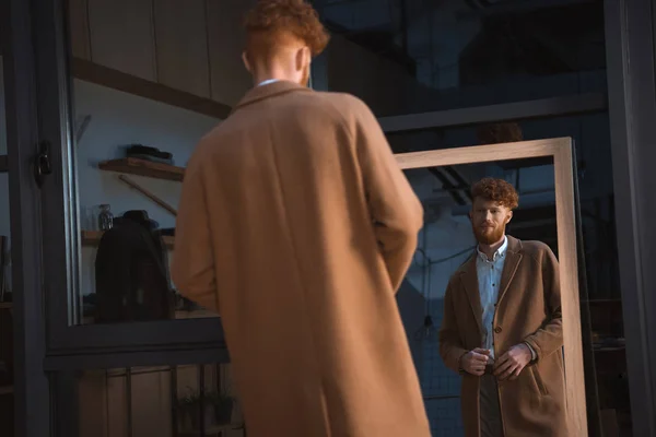 Back view of young man wearing overcoat and looking at mirror in boutique — Stock Photo