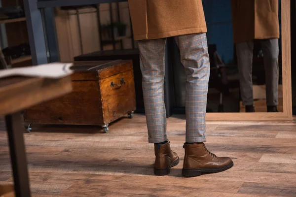 Seção baixa do homem em botas de couro e sobretudo de pé perto do espelho na boutique — Fotografia de Stock