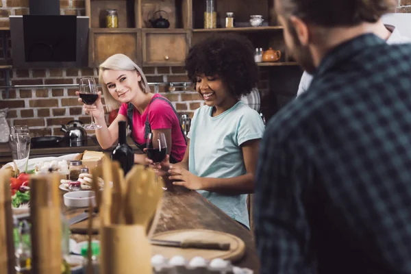 Meninas multiétnicas com vinho olhando para os amigos na cozinha — Fotografia de Stock