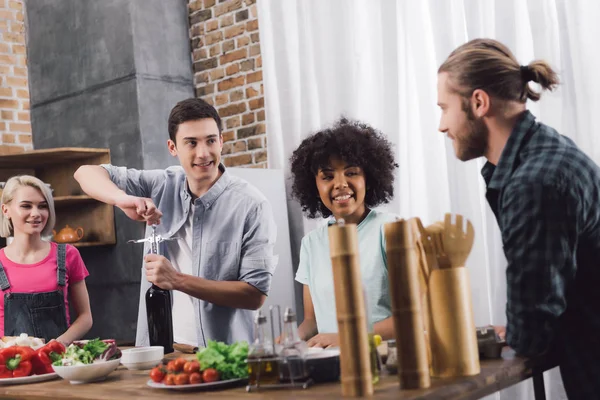 Uomo aprendo bottiglia di vino con cavatappi e parlando con amici multietnici — Foto stock