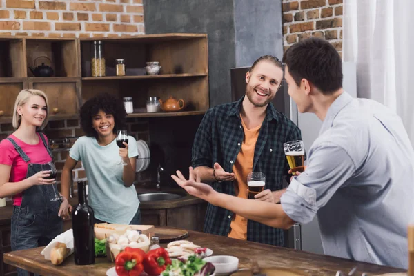 Улыбающиеся многонациональные друзья разговаривают на кухне с пивом и вином — стоковое фото