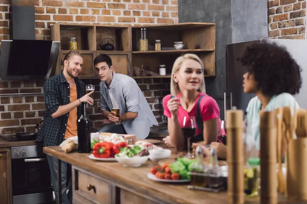 Multiethnic girls talking in kitchen with wine and male friends looking at them — Stock Photo